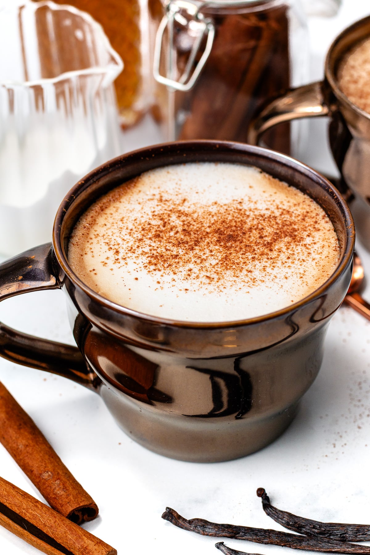 A brown ceramic mug filled with a frothy, spiced latte topped with ground cinnamon, surrounded by cinnamon sticks and vanilla beans on a white surface.