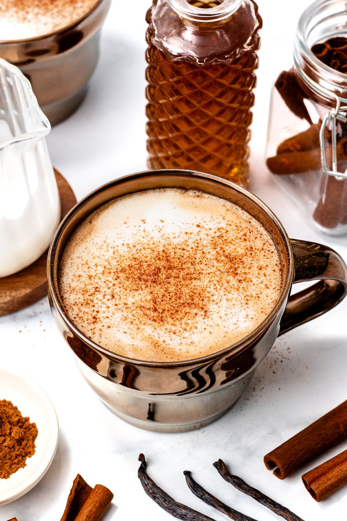 A mug of frothy latte topped with cinnamon sits on a white surface, surrounded by cinnamon sticks, vanilla beans, a glass bottle of syrup, and a small pitcher of milk.