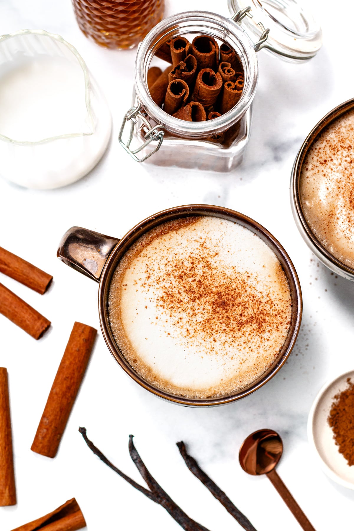 A cup of frothy coffee topped with cinnamon sits on a white surface, surrounded by cinnamon sticks, a jar of cinnamon, a milk pitcher, and a small bowl with a spoon.