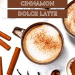 Top-down view of a cinnamon dolce latte with cinnamon sticks, vanilla beans, a spoon of sugar, and a second mug. Text overlay reads "Copycat Cinnamon Dolce Latte.
