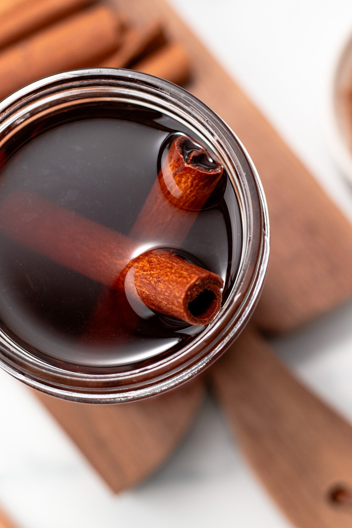 A close-up of a glass jar filled with dark liquid and two cinnamon sticks, placed on a wooden surface.