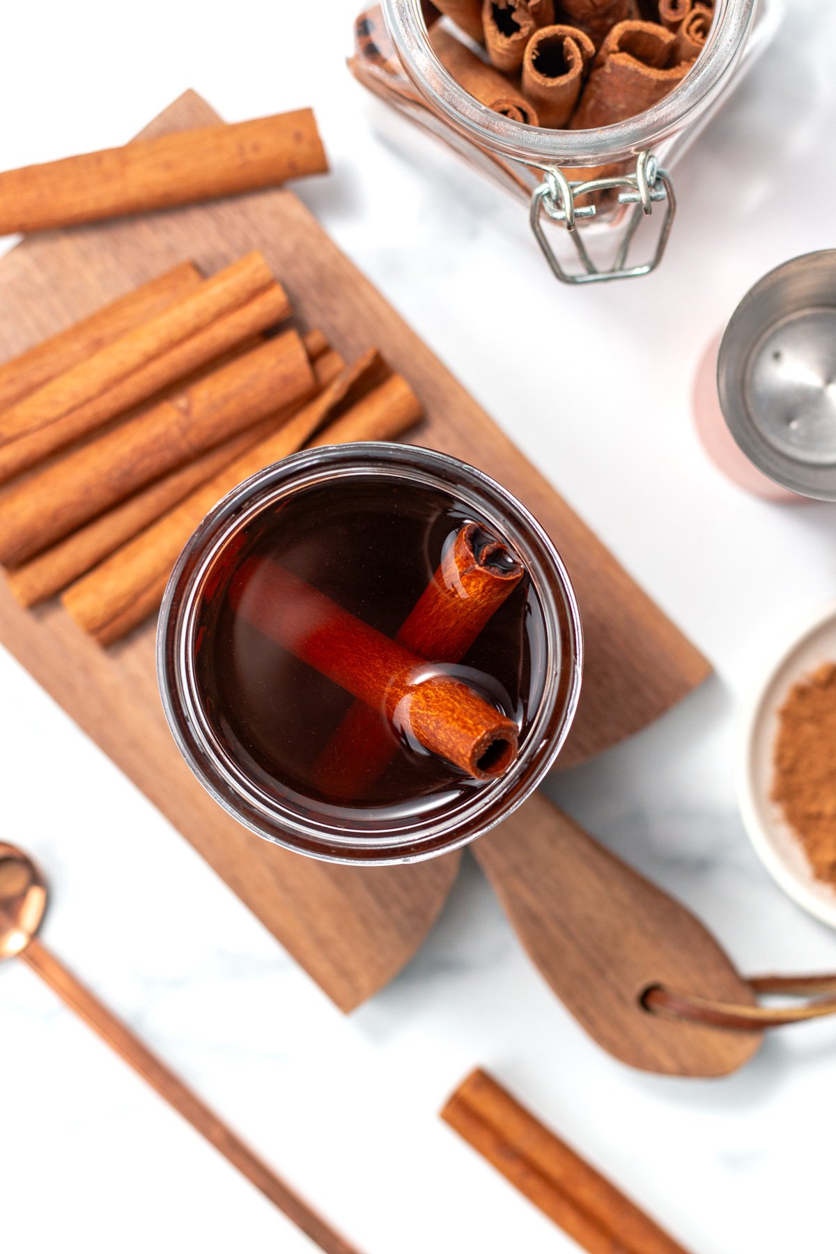 A glass of dark liquid with two cinnamon sticks inside, surrounded by a jar of cinnamon sticks, a wooden board, and a small bowl of ground cinnamon.
