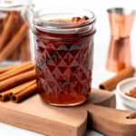 A glass jar filled with a dark beverage and cinnamon sticks, placed on a wooden board, surrounded by cinnamon sticks, a copper measuring cup, and a spoon.