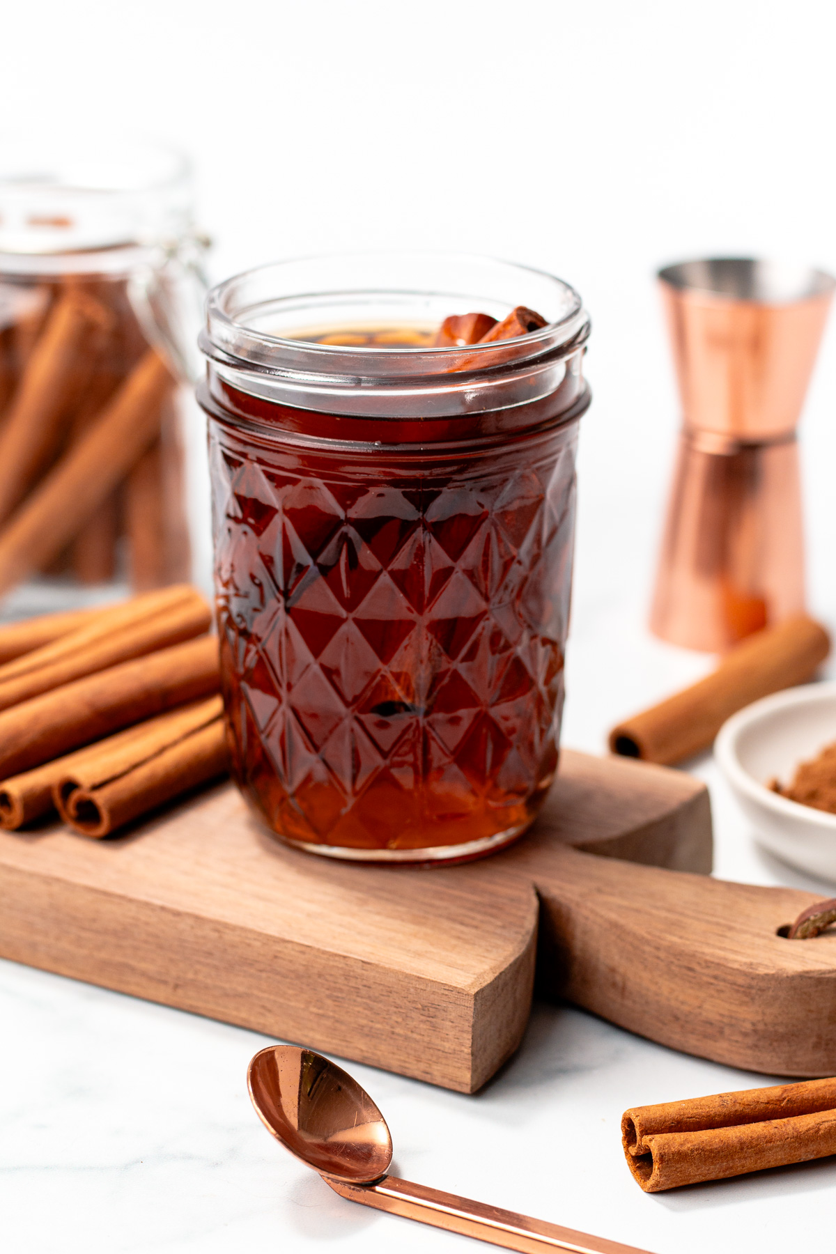 A glass jar filled with a dark beverage and cinnamon sticks, placed on a wooden board, surrounded by cinnamon sticks, a copper measuring cup, and a spoon.
