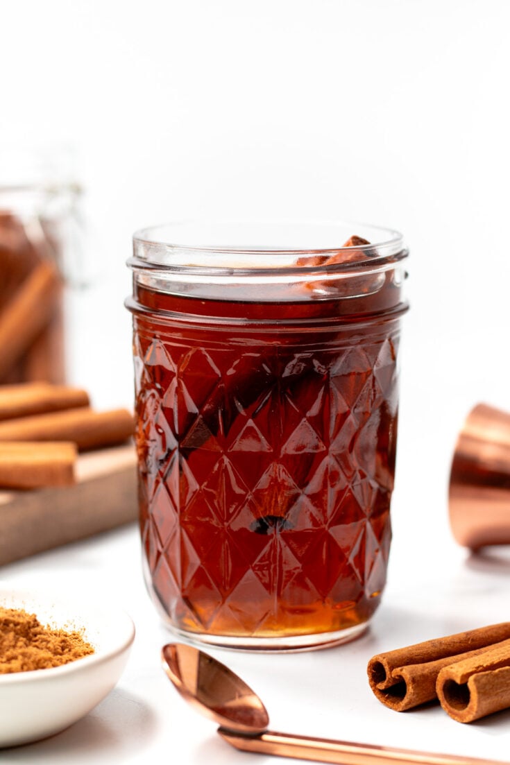 A glass jar filled with dark brown liquid, garnished with cinnamon sticks. Nearby are more cinnamon sticks, ground cinnamon in a dish, and a copper measuring cup.
