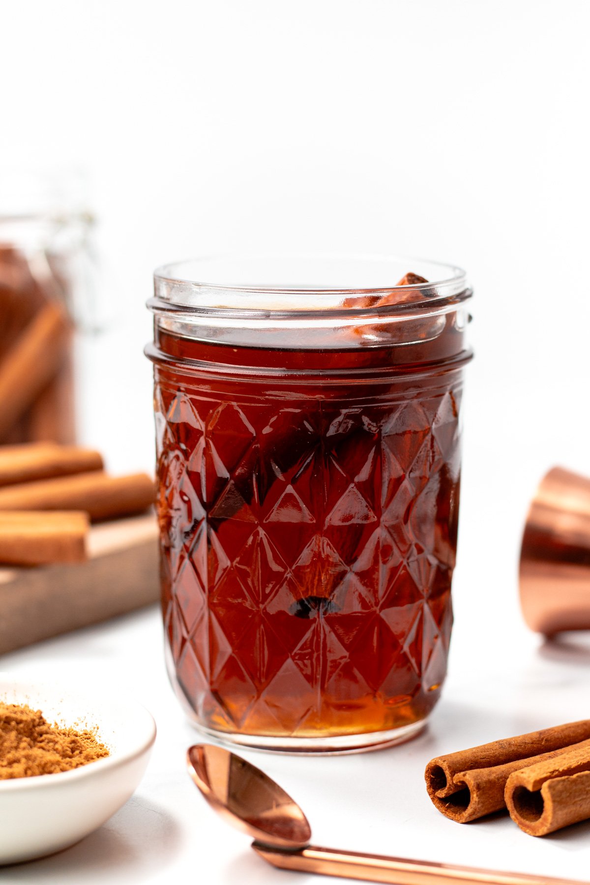 A glass jar filled with dark brown liquid, garnished with cinnamon sticks. Nearby are more cinnamon sticks, ground cinnamon in a dish, and a copper measuring cup.
