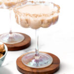 A creamy Cinnamon Toast Crunch cocktail in a wide glass with a crumb-coated rim, set on a wooden coaster, with cereal pieces nearby and another similar cocktail in the background.