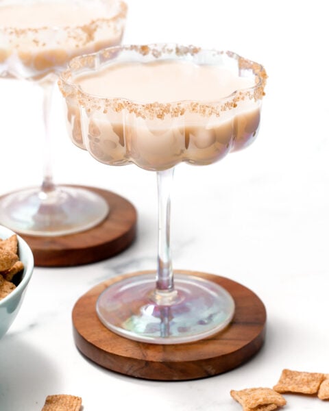 A creamy Cinnamon Toast Crunch cocktail in a wide glass with a crumb-coated rim, set on a wooden coaster, with cereal pieces nearby and another similar cocktail in the background.