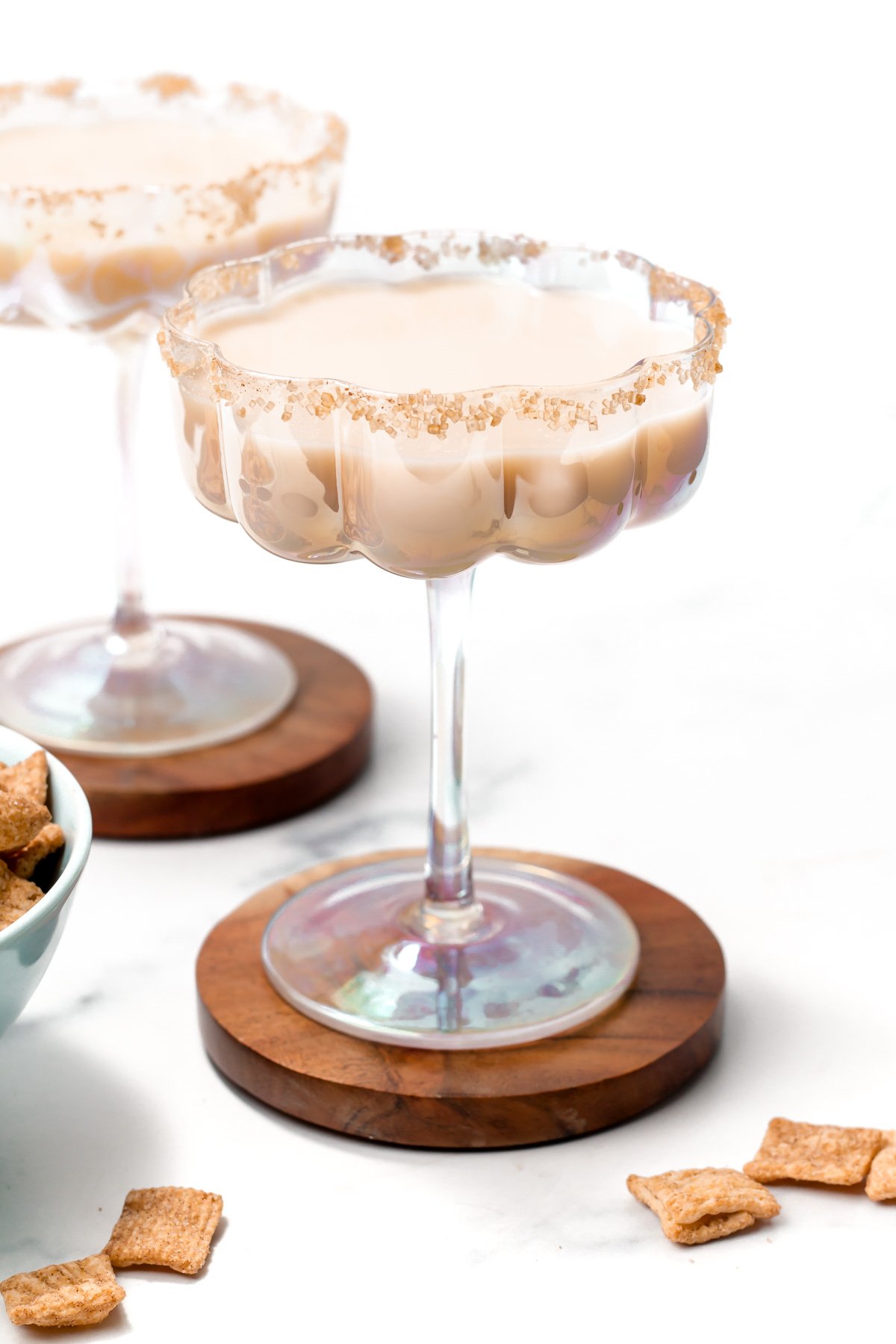 A creamy Cinnamon Toast Crunch cocktail in a wide glass with a crumb-coated rim, set on a wooden coaster, with cereal pieces nearby and another similar cocktail in the background.