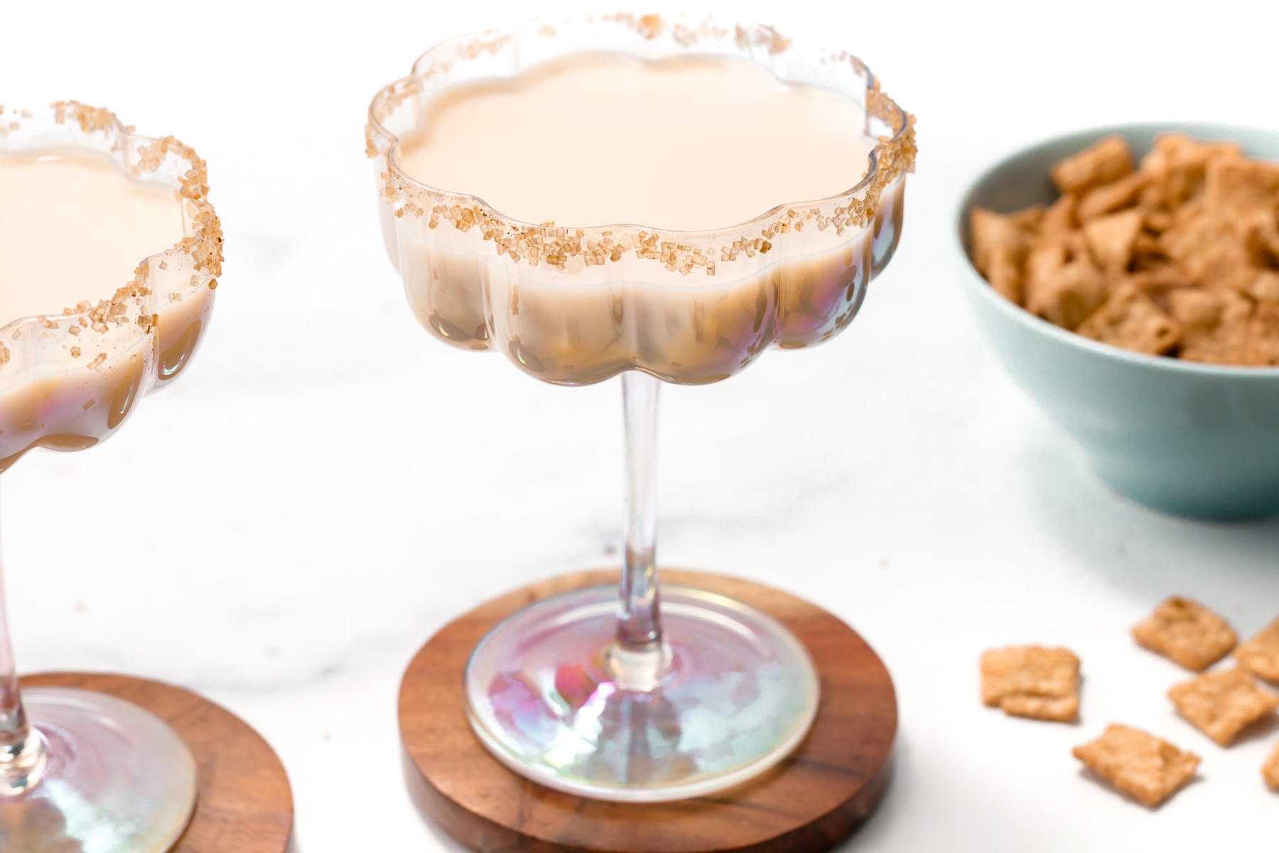 A Cinnamon Toast Crunch Cocktail in a decorative glass with a brown sugar rim sits on a wooden coaster; a bowl of cereal is in the background.