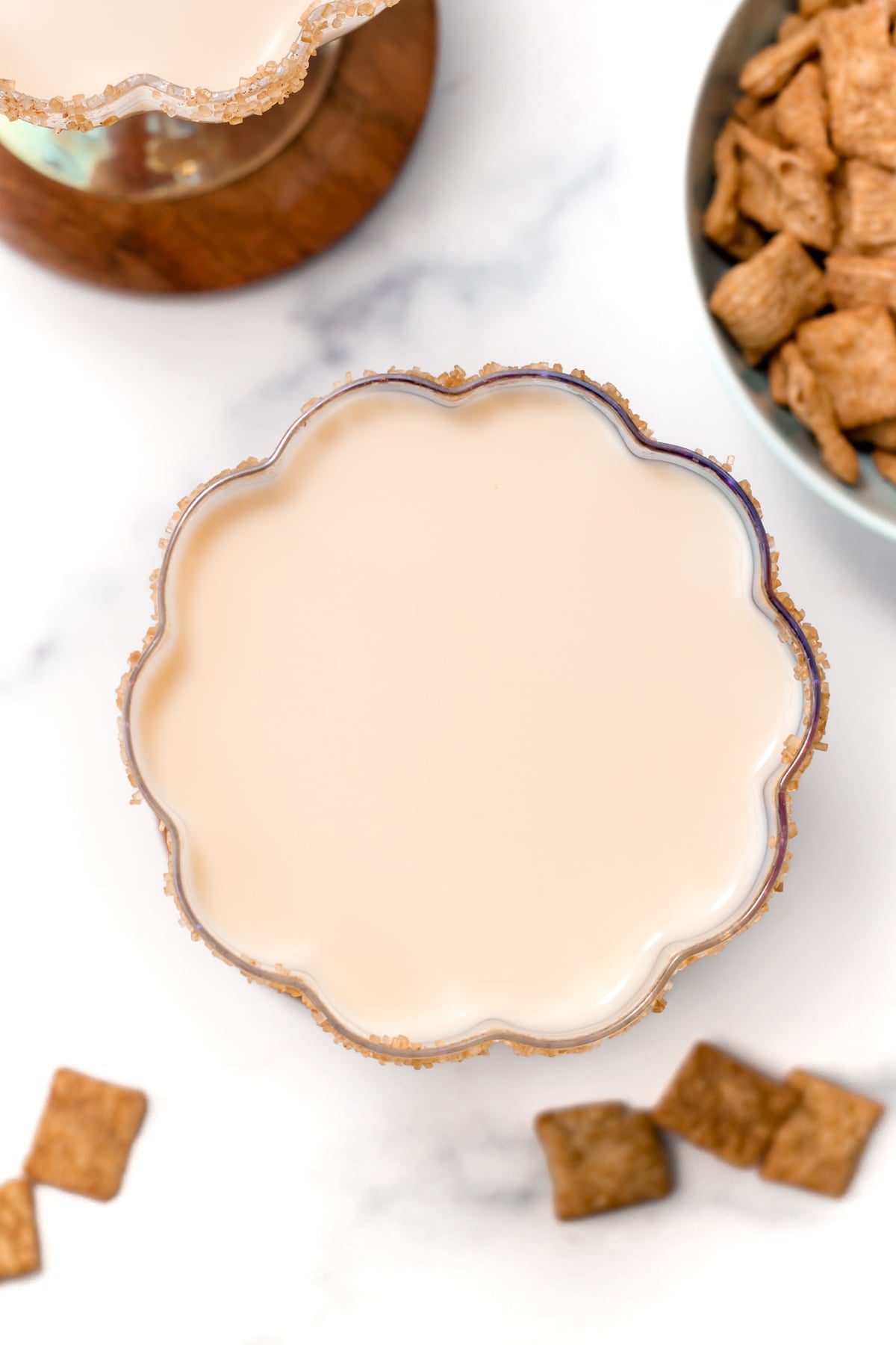A scalloped glass filled with a creamy beige Cinnamon Toast Crunch cocktail, rimmed with brown sugar, sits on a white surface next to a bowl of cereal squares and scattered cereal pieces.
