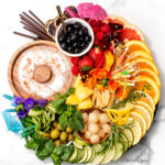 A round platter with sliced citrus, cucumbers, fresh herbs, olives, fruit, lychee, chips, chocolate sticks, black cherries, and cocktail umbrellas on a white marble surface.