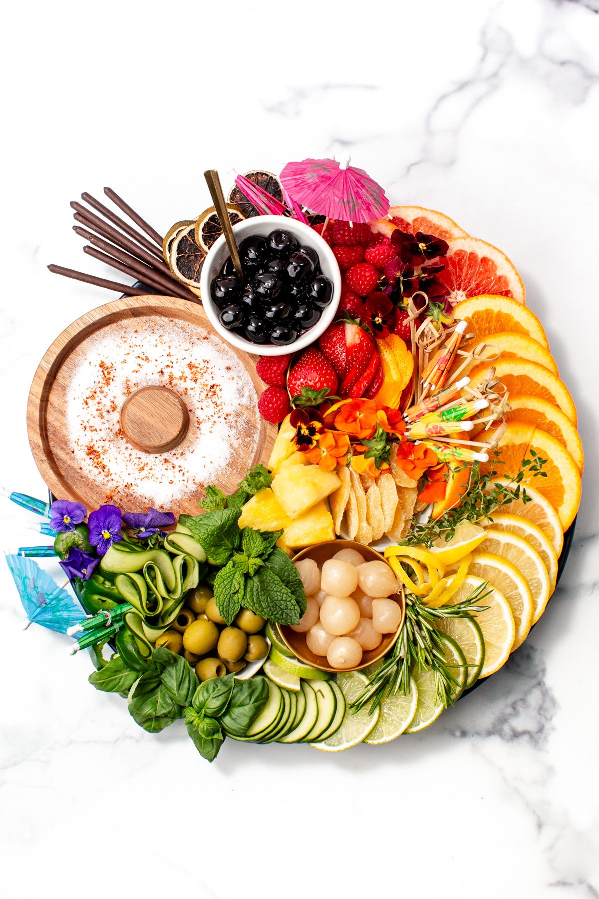 A round platter with sliced citrus, cucumbers, fresh herbs, olives, fruit, lychee, chips, chocolate sticks, black cherries, and cocktail umbrellas on a white marble surface.