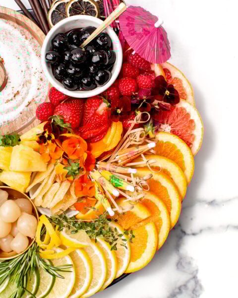 A platter with sliced oranges, lemons, grapefruits, pineapple, strawberries, raspberries, cherries, lychees, herbs, edible flowers, drink umbrellas, and cocktail sticks on a marble surface.