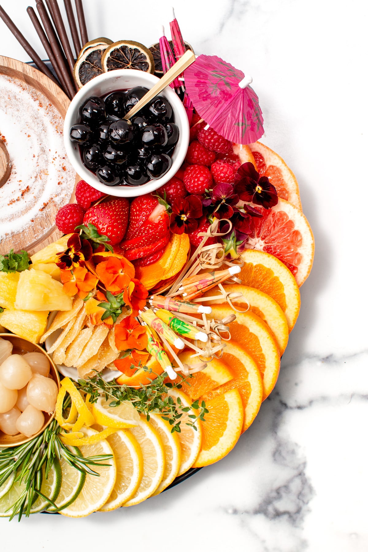 A platter with sliced oranges, lemons, grapefruits, pineapple, strawberries, raspberries, cherries, lychees, herbs, edible flowers, drink umbrellas, and cocktail sticks on a marble surface.