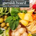 A platter with green olives, fresh mint, pineapple, pearl onions, cucumber slices, citrus peel, chips, and edible flowers, labeled as an easy cocktail garnish board.