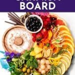 A platter with assorted cocktail garnishes including citrus slices, berries, herbs, candy, and flowers, arranged around a dish of salt; text reads "Cocktail Garnish Board.