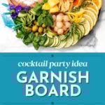 A colorful cocktail garnish board with fruits, herbs, flowers, pickled items, cheese, and dip is artfully arranged on a round platter—perfect as a festive cocktail party idea.