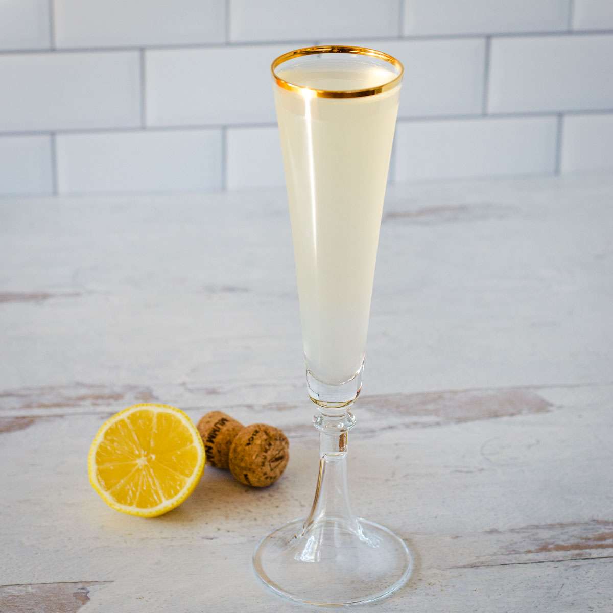 A champagne flute with a pale drink, garnished with a gold rim, sits on a light surface next to a halved lemon and a champagne cork.