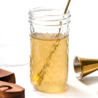 A glass jar filled with a light yellow liquid, a metal spoon inside, ginger root and slices on a wooden board, a metal jigger, and a small bowl with ginger pieces nearby.