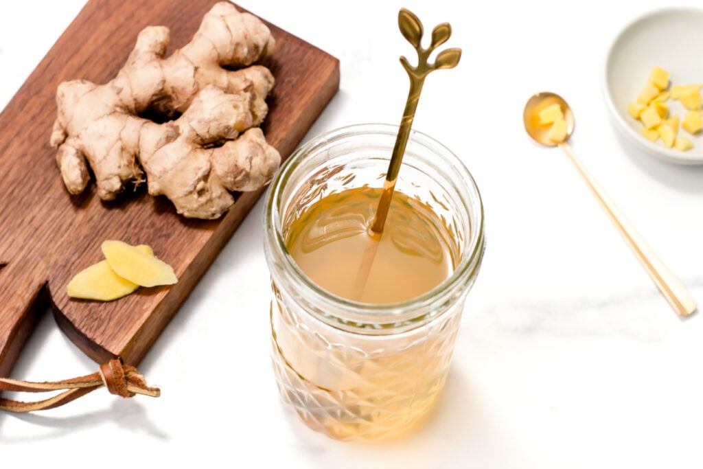 A glass jar of ginger tea with a gold spoon, fresh ginger root on a wooden board, and a small bowl of ginger pieces on a white surface.