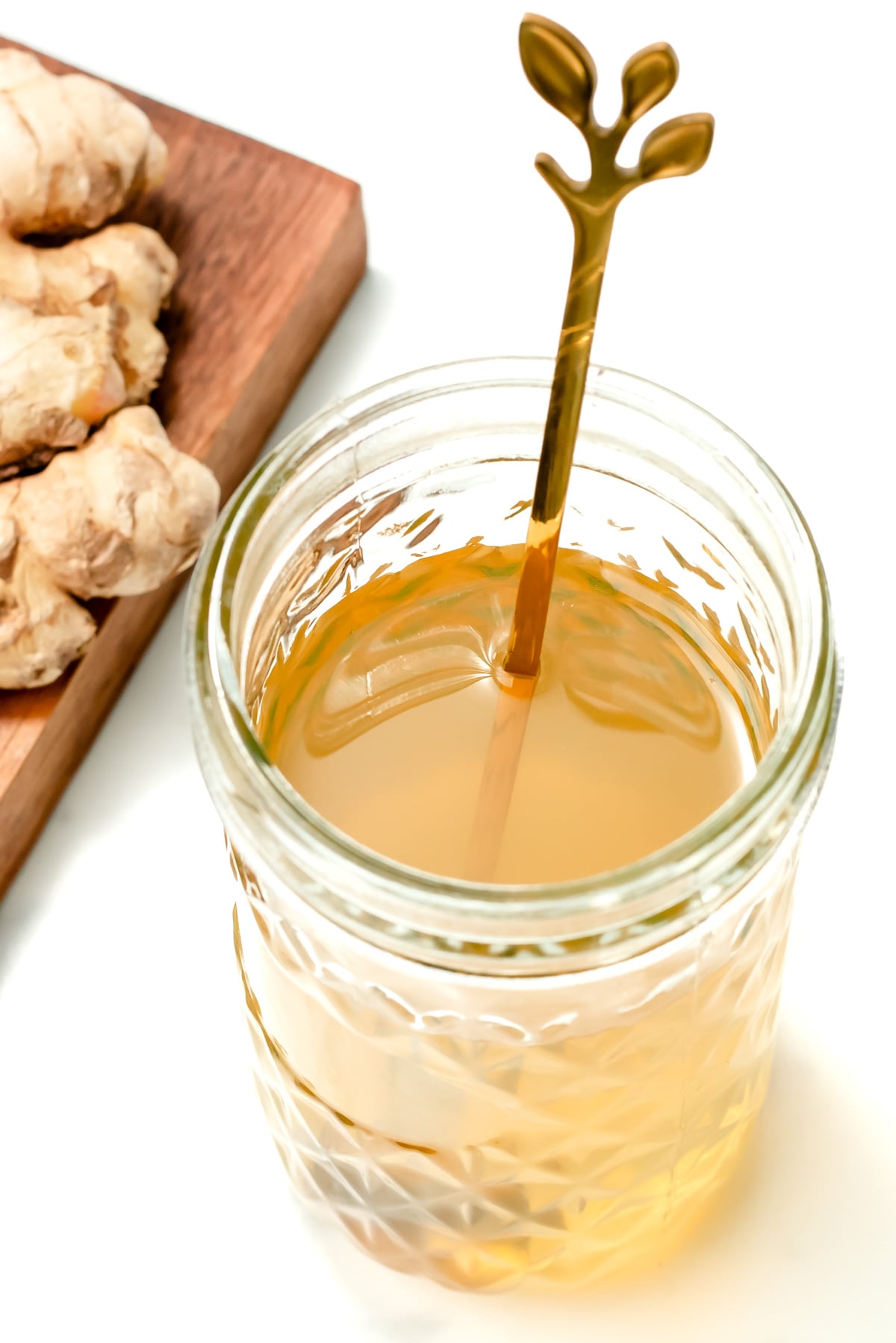A glass jar filled with a light brown liquid and a decorative gold spoon, next to a wooden board with sliced ginger.