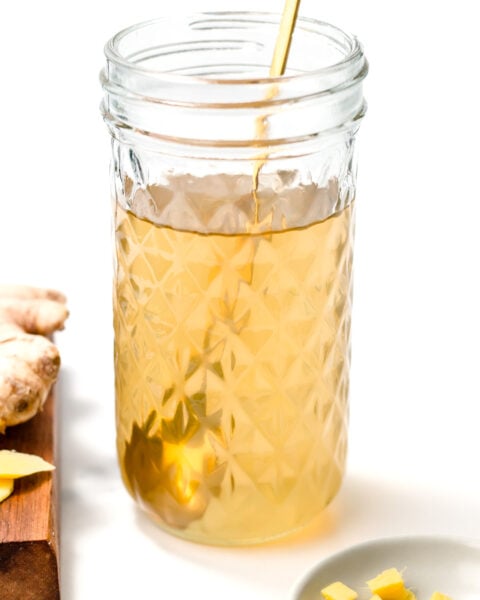 A clear glass jar filled with light yellow liquid, a gold spoon inside, ginger root on a wooden board, and chopped ginger on a small plate nearby.
