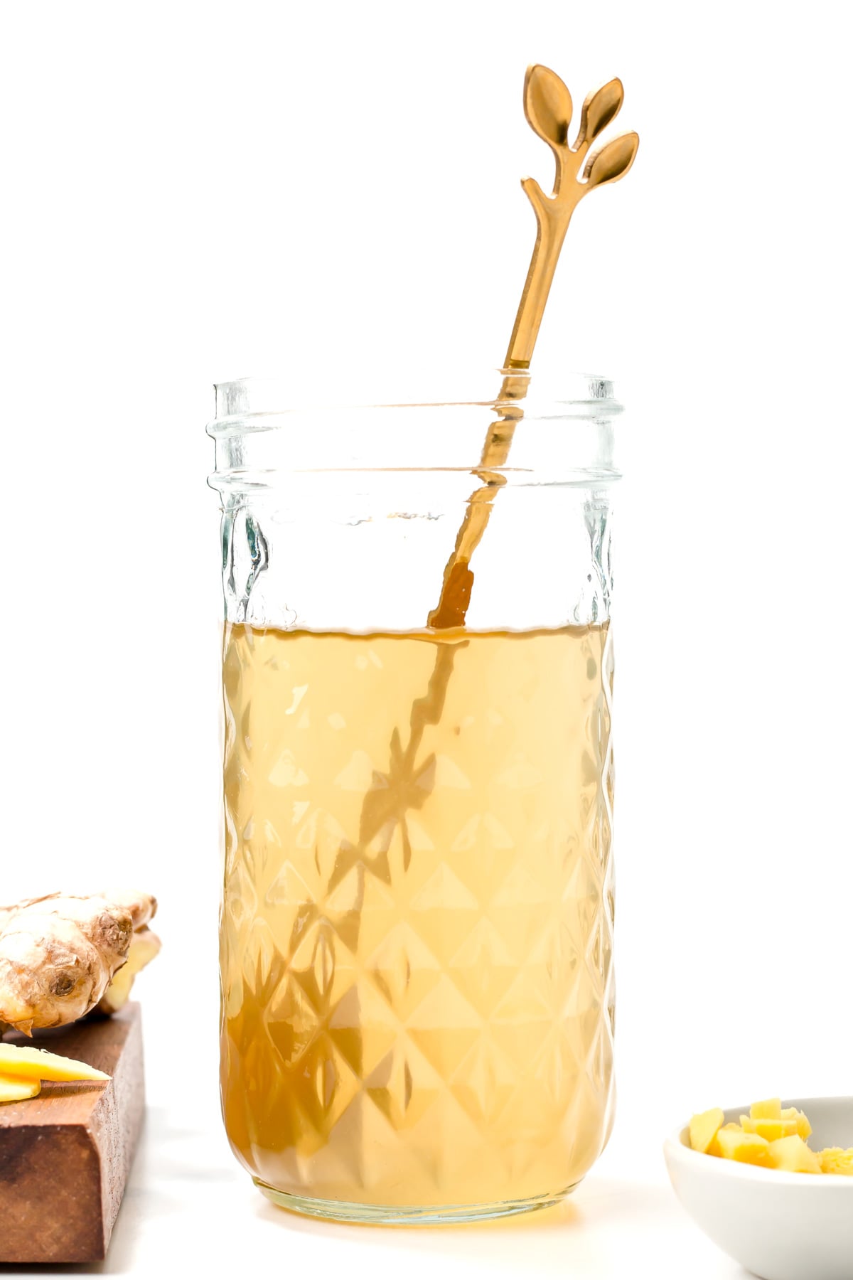 A mason jar filled with light yellow liquid, a gold spoon inside, and ginger pieces and a bowl of diced fruit beside it on a white background.
