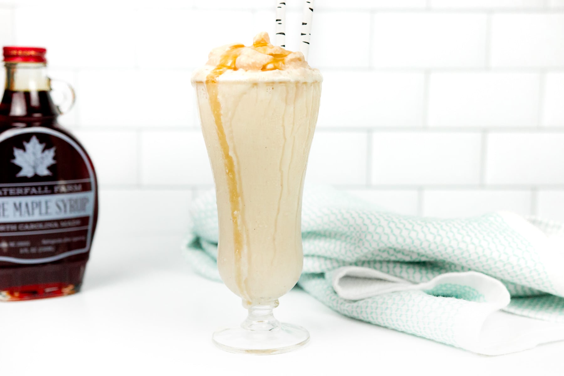 A glass of caramel milkshake with whipped cream and caramel drizzle, served with two striped straws. A bottle of maple syrup and a folded towel are in the background.