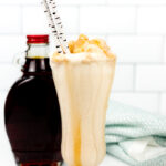 A glass of Maple Bourbon Milkshake with caramel drizzle, topped with whipped cream and two straws, sits next to a bottle of syrup on a white surface.