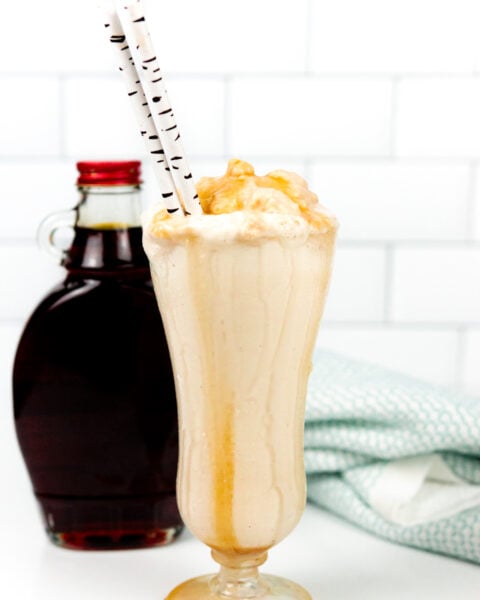 A tall glass of caramel milkshake with whipped topping and caramel drizzle, two straws, and a syrup bottle in the background.