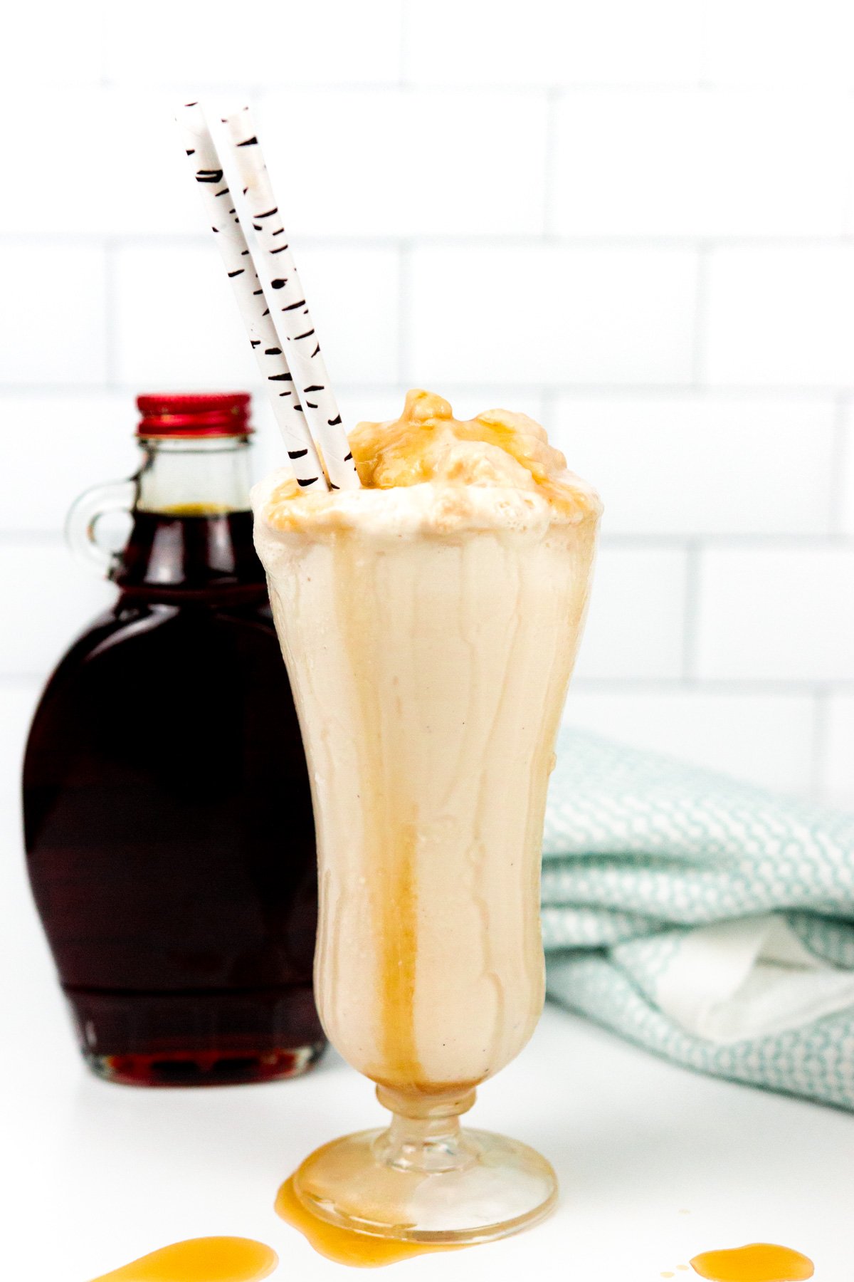 A tall glass of caramel milkshake with whipped topping and caramel drizzle, two straws, and a syrup bottle in the background.