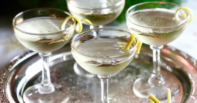 Four coupe glasses filled with a clear cocktail, each garnished with a lemon twist, are arranged on a reflective silver tray.