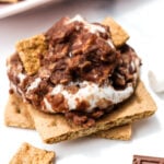 A close-up of a s'mores treat with a graham cracker base, melted marshmallow, chocolate, and cereal pieces on top, surrounded by small marshmallows and chocolate squares.