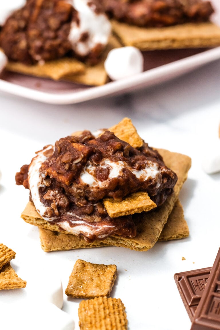 A close-up of a s’more made with graham crackers, chocolate, marshmallow, and a cereal topping, with extra cereal and chocolate pieces scattered around.