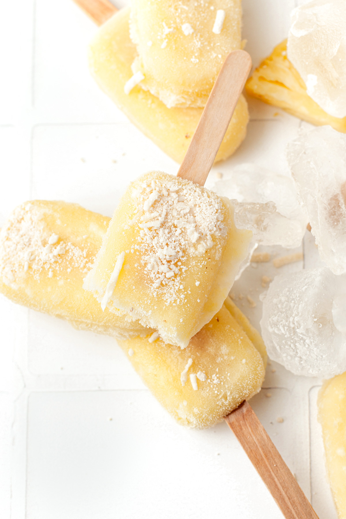 Three yellow popsicles with shredded coconut and ice cubes on a white surface, arranged in a casual stack.