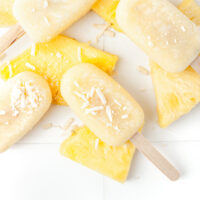 Four pineapple popsicles with coconut flakes are arranged on top of several slices of fresh pineapple on a white surface.