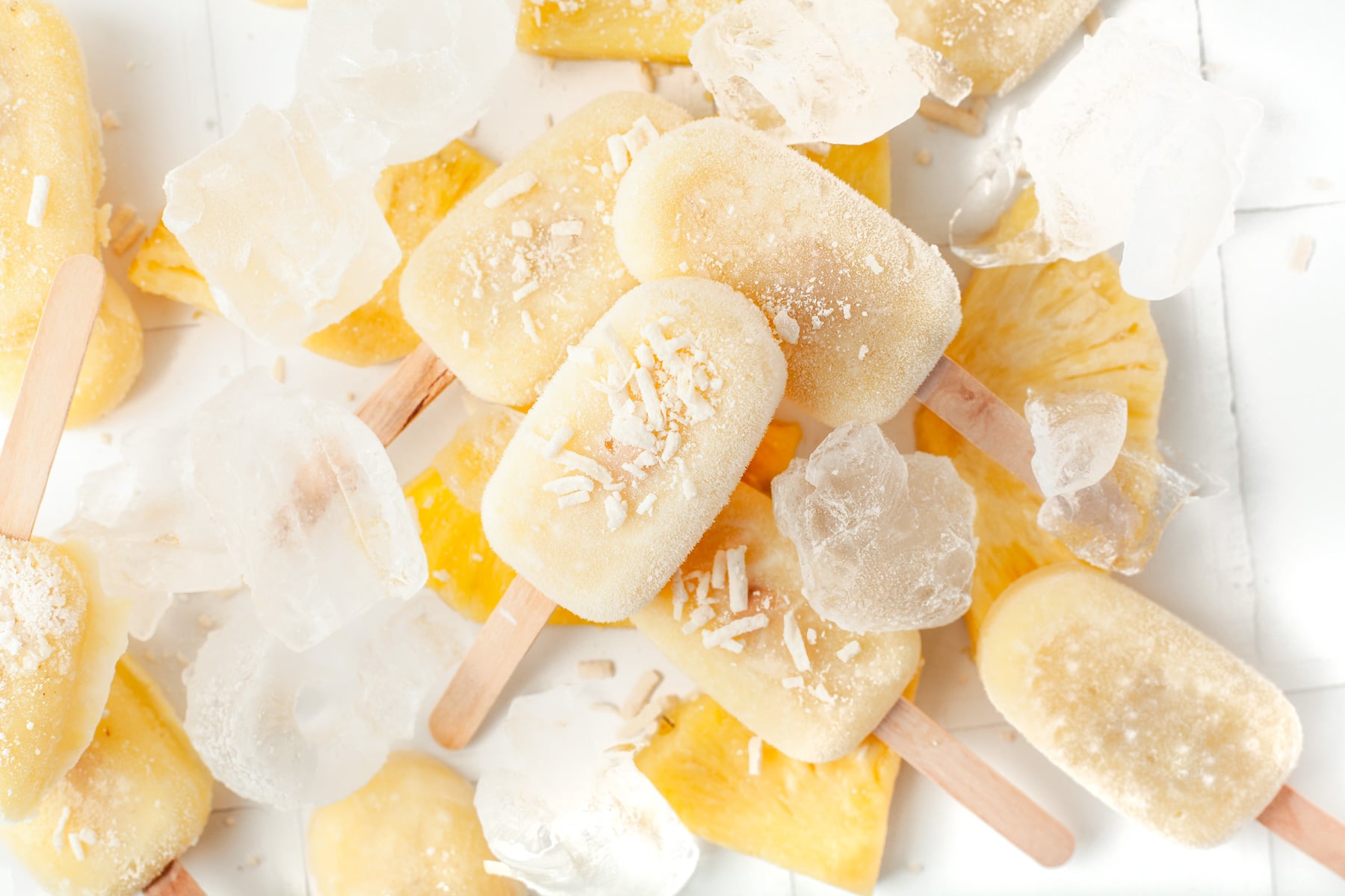 Several yellow popsicles with shredded coconut on top are arranged with ice cubes and pineapple slices on a white surface.