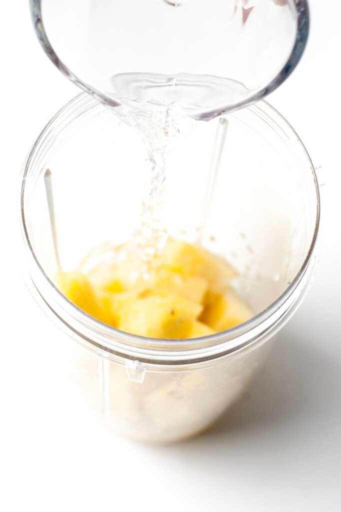 Water being poured into a blender cup containing pineapple chunks, set against a white background.