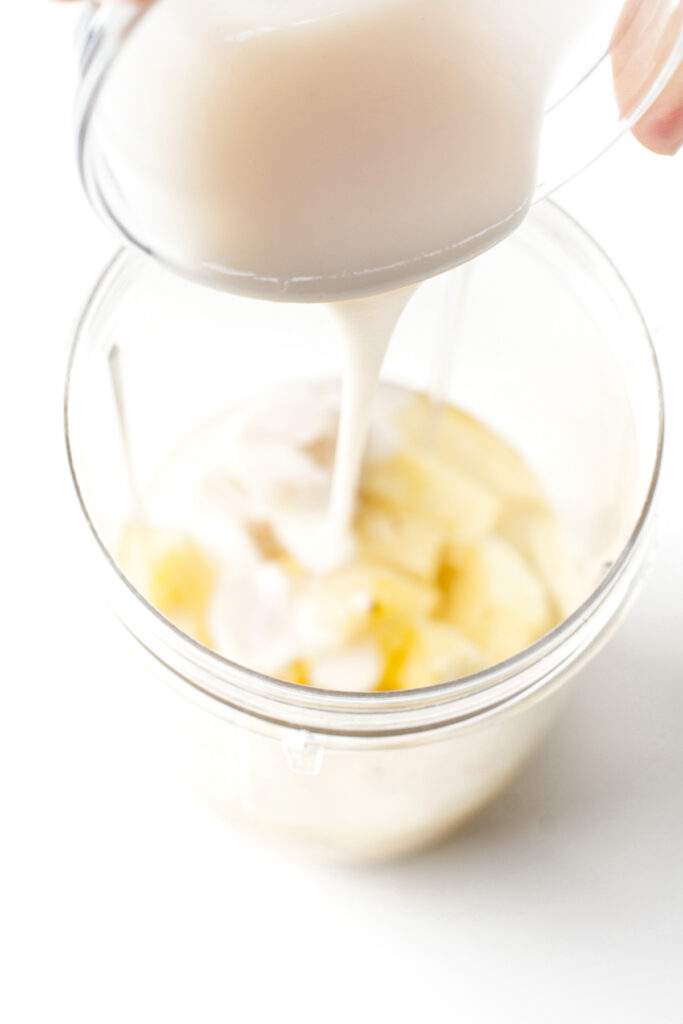 A creamy liquid is being poured into a blender cup containing sliced bananas.