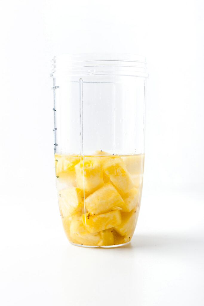 A clear blender cup filled with pineapple chunks and liquid, perfect for making refreshing pi&ntilde;a colada popsicles, sits on a white surface with a white background.