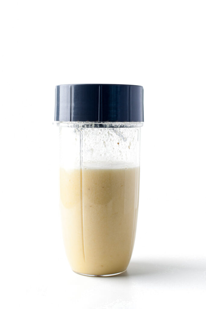 A clear plastic blender cup filled with a light yellow smoothie, reminiscent of pi&ntilde;a colada popsicles, sealed with a dark blue lid and set against a white background.