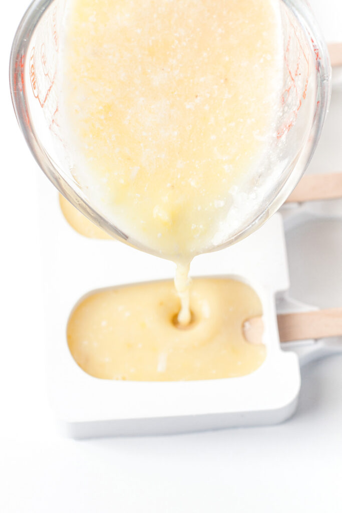A measuring cup pours yellow batter into a white popsicle mold with wooden sticks.