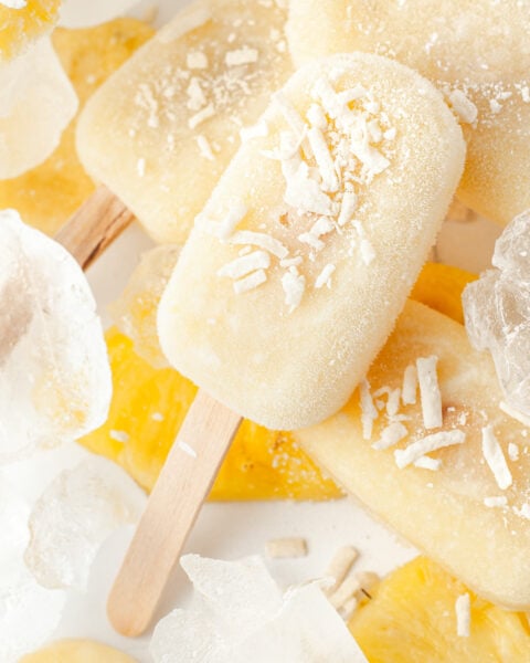 Close-up of yellow popsicles with shredded coconut on top, surrounded by ice cubes and pineapple slices.