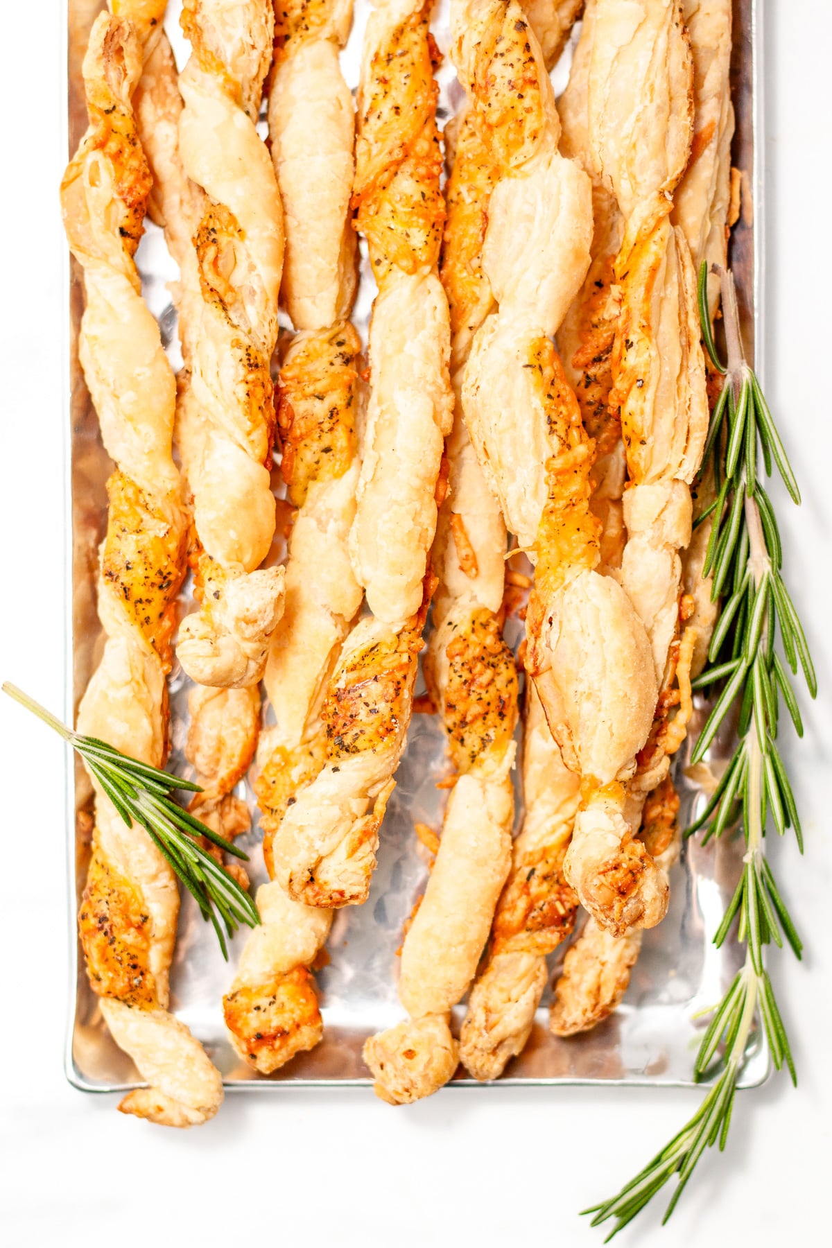 A tray of twisted puff pastry cheese straws garnished with sprigs of fresh rosemary.