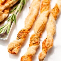 Four twisted puff pastry sticks seasoned with herbs and cheese, arranged on a white surface next to a sprig of fresh rosemary.