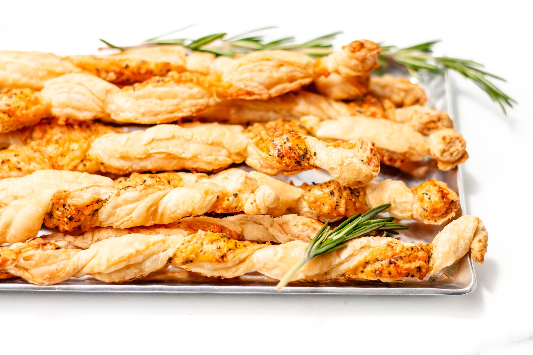 A tray of golden, twisted cheese straws garnished with fresh rosemary on a white background.