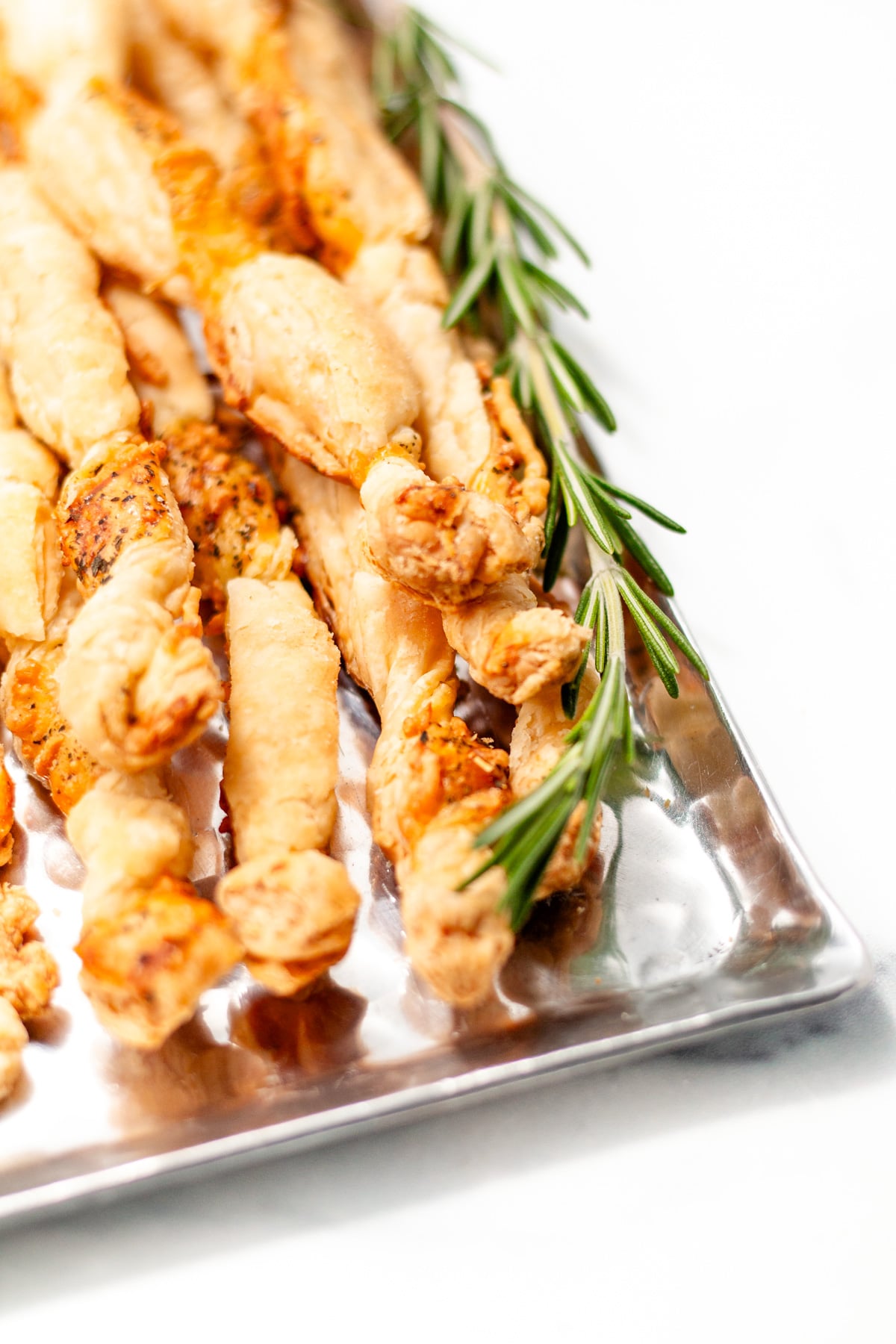 A silver tray with twisted breadsticks garnished with fresh rosemary on a white background.