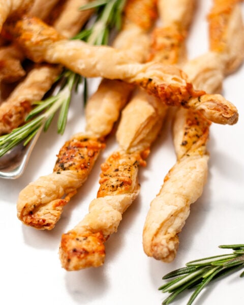 Close-up of twisted puff pastry breadsticks sprinkled with herbs and cheese, garnished with fresh rosemary sprigs on a white surface.