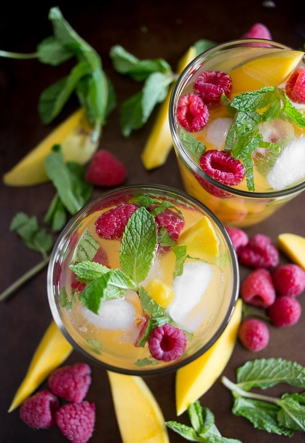 Two glasses of iced fruit drinks garnished with fresh mint, raspberries, and mango slices, surrounded by additional mint leaves, mango slices, and raspberries on a dark surface.